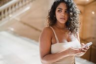Crop concentrated young Hispanic female with dark curly hair in casual clothes messaging on mobile phone and looking away thoughtfully while waiting for meeting