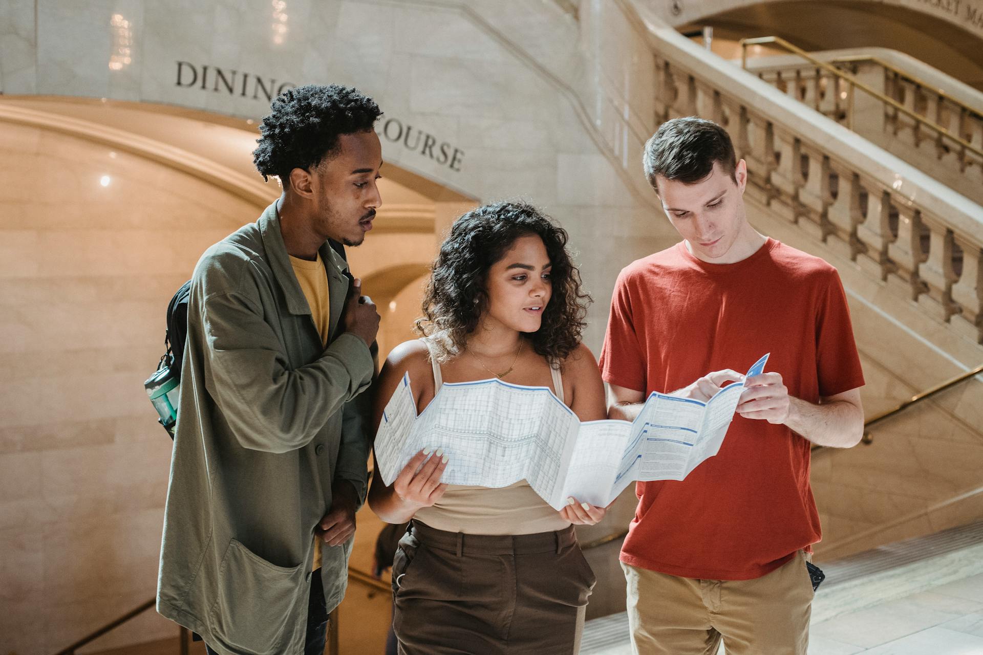 Focused young man pointing at map while searching for route with multiracial friends in Grand Central Terminal during trip in New York