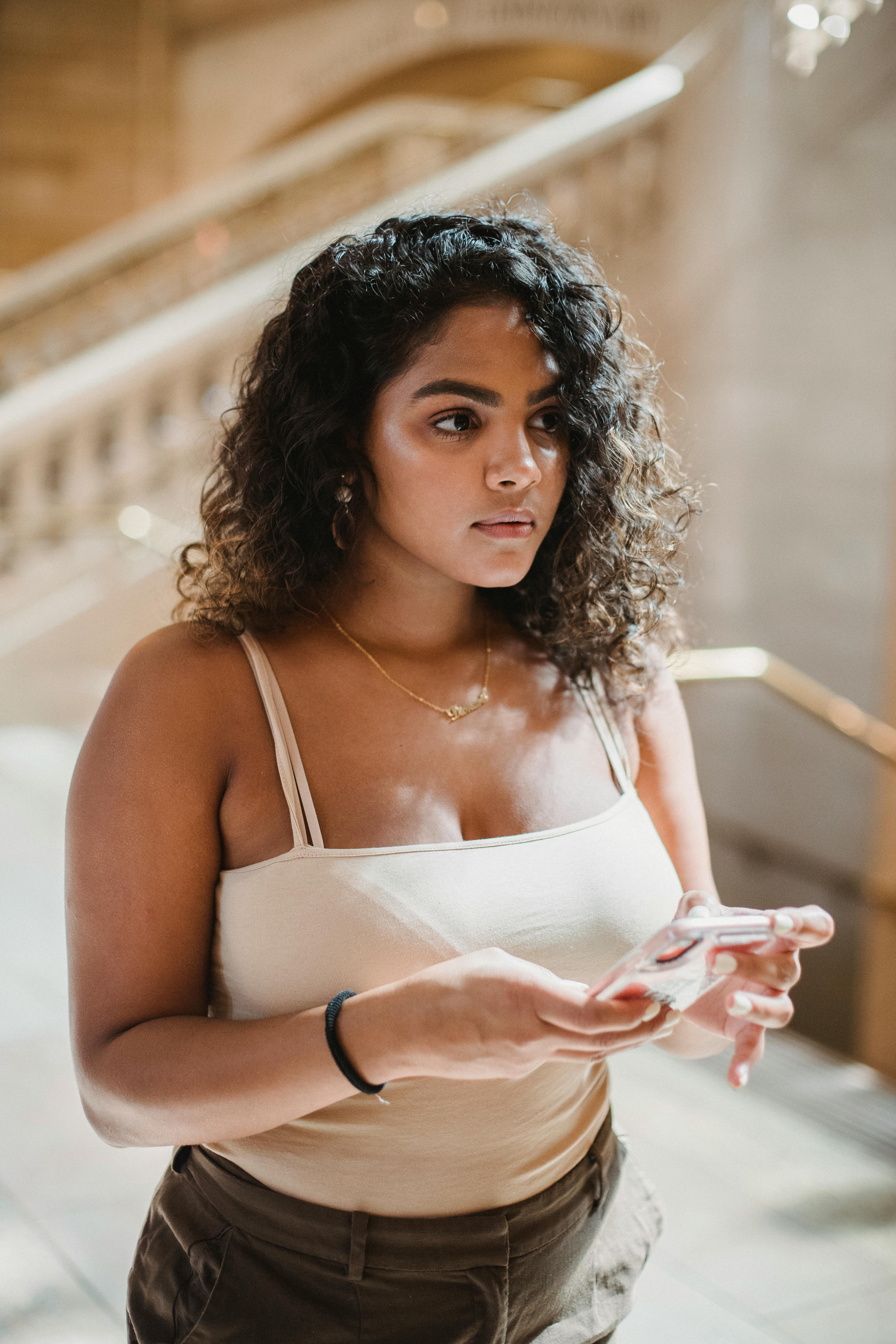 pensive young ethnic female messaging on smartphone and looking away