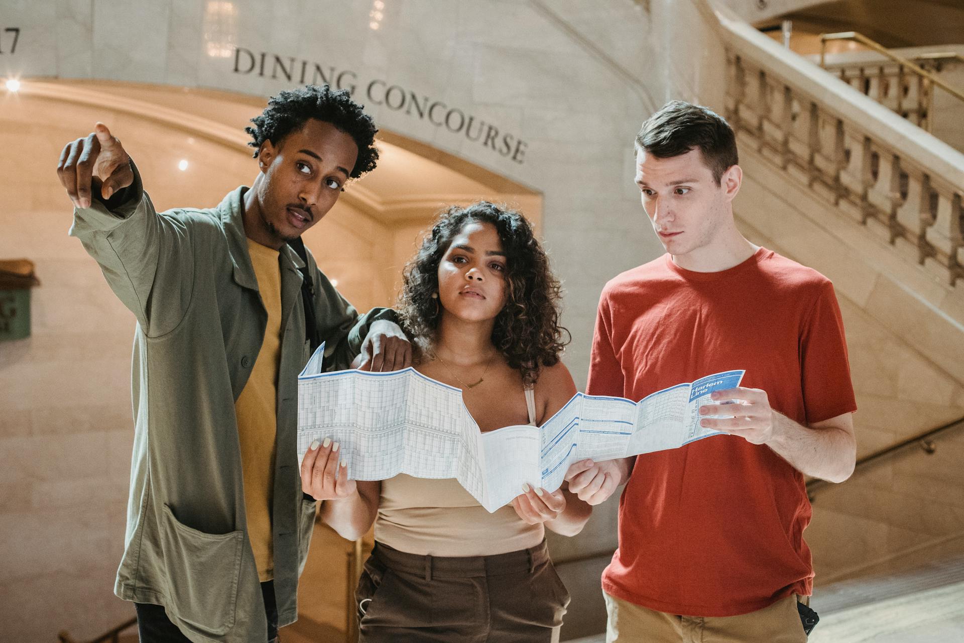 Young African American male tourist pointing away while searching for direction with diverse fiends standing in railway station terminal with paper map in hands