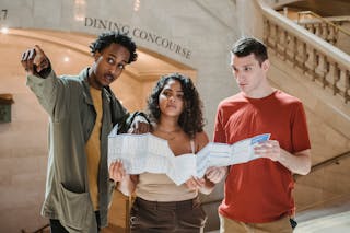 Young African American male tourist pointing away while searching for direction with diverse fiends standing in railway station terminal with paper map in hands
