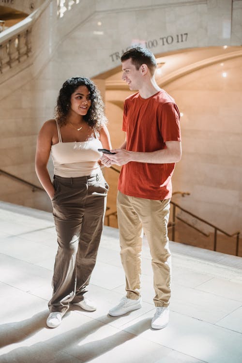 Full body of positive young multiracial friends in stylish outfits smiling and chatting while using smartphone in aged building hallway