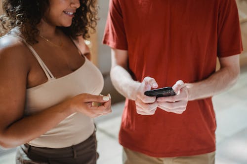 Free Crop unrecognizable man in casual clothes messaging on smartphone while standing near smiling Hispanic girlfriend Stock Photo