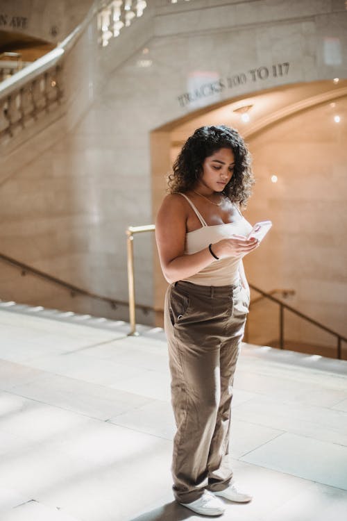 Side view of young focused ethnic lady with curly dark hair in casual clothes standing in Grand Central Terminal and using mobile phone