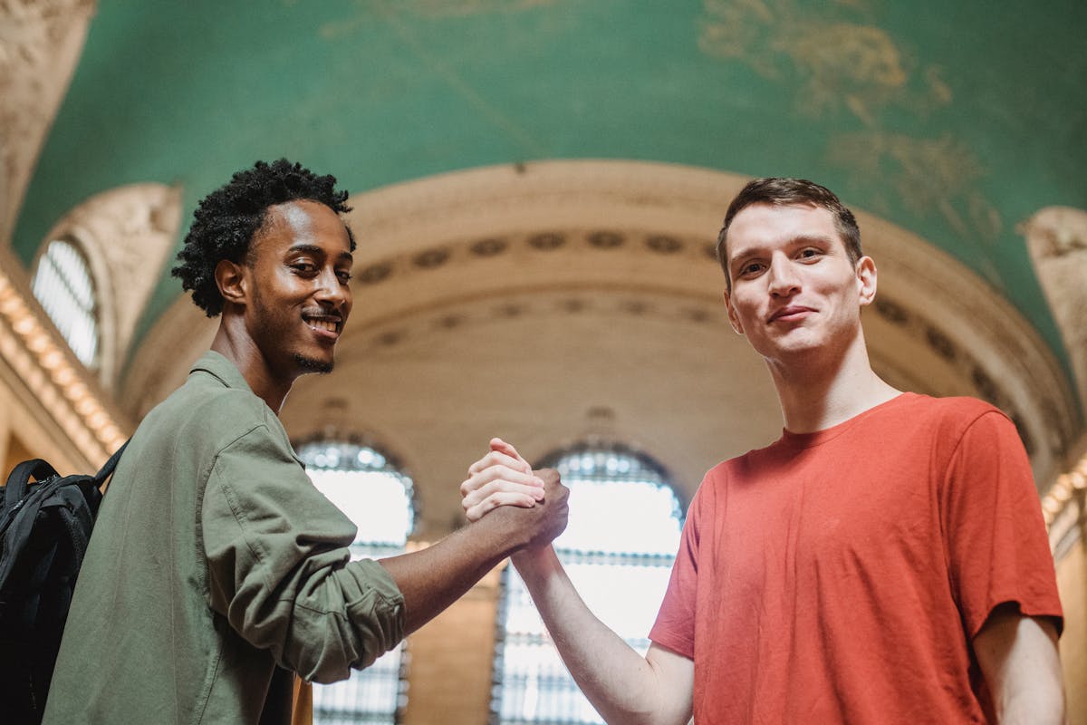 From below of positive young multiethnic male friends in casual clothes smiling and looking at camera while shaking hands in spacious aged arched building