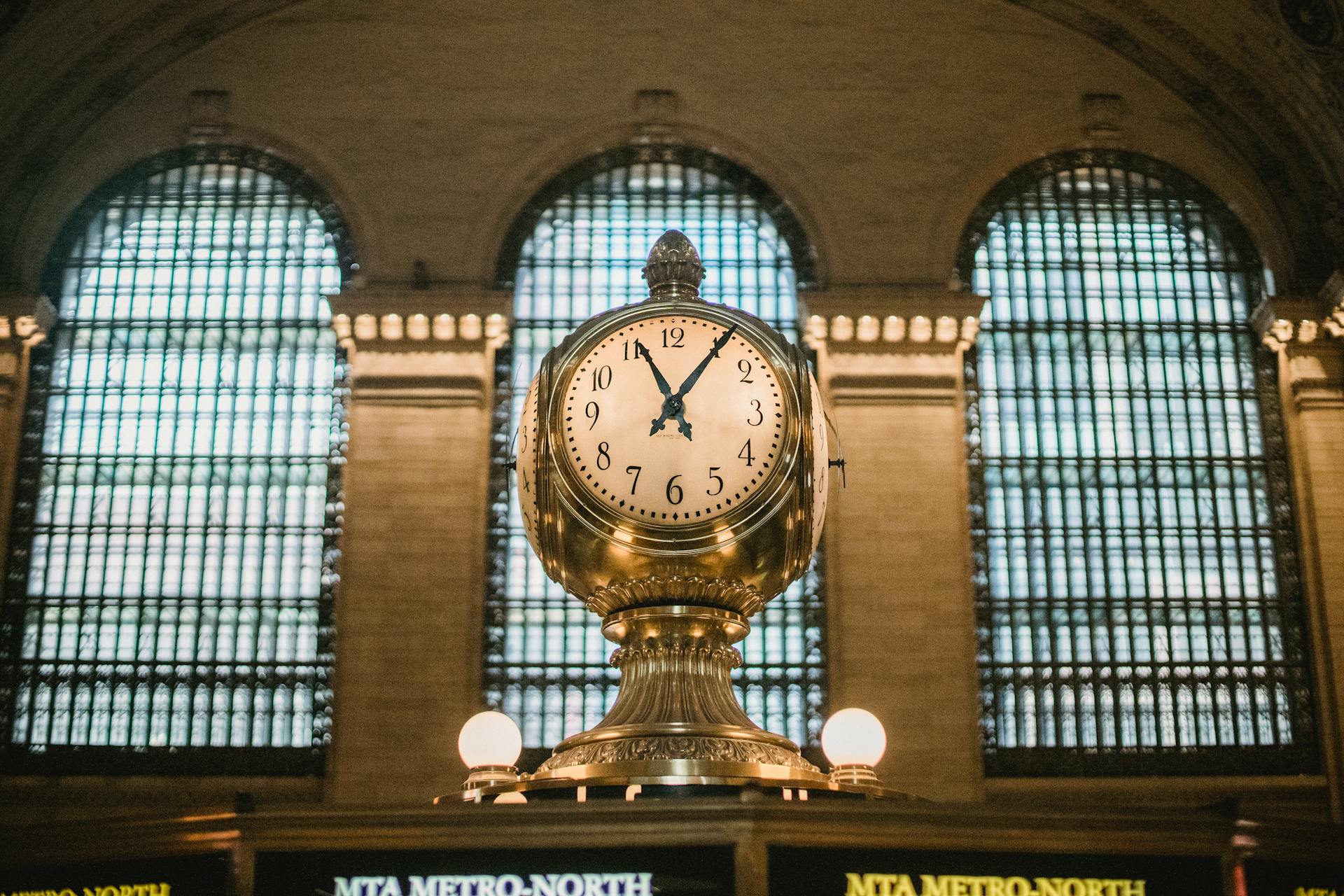 From below of aged retro golden clock placed atop information booth of historic Grand Central Terminal with arched windows