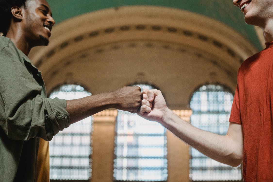 Free From below of crop multiethnic friends in casual clothes giving fist bump against arched wall and windows in aged hallway of Grand Central Terminal placed in New York City Stock Photo