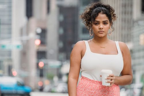 Free Confident Hispanic woman with curly hair in trendy clothes standing with white cup of takeaway drink on on city street and looking at camera in daytime Stock Photo