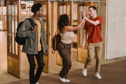 Full body of multiracial group of students looking at each other and giving high five while strolling through doorway in corridor of university building