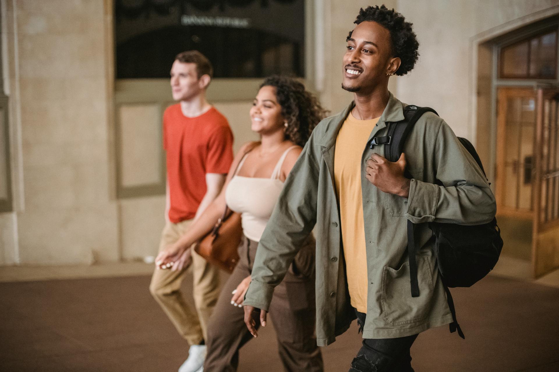 Positive African American man with male friend and Hispanic woman strolling in hallway of university building in campus on blurred background