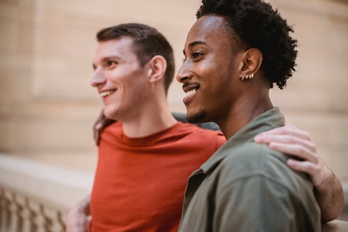 Side view of cheerful multiracial male friends hugging and looking into distance while standing near stone handrail on blurred background