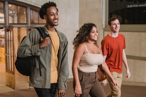 Free Cheerful multiracial group of friends with backpacks in casual clothes strolling together in corridor of university building with doorway on background Stock Photo