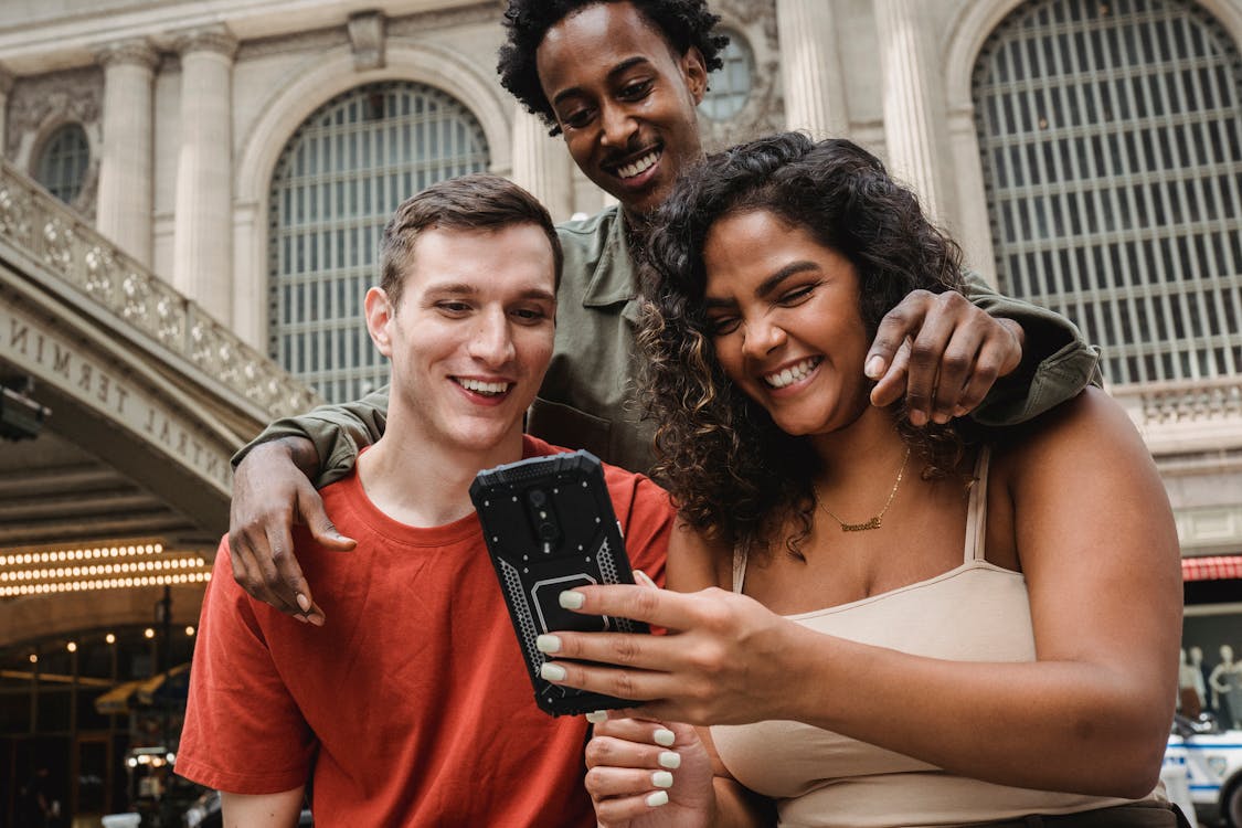 Free From below of delighted multiracial group of friends hugging and looking at screen of cellphone while spending time on street against historic building Stock Photo