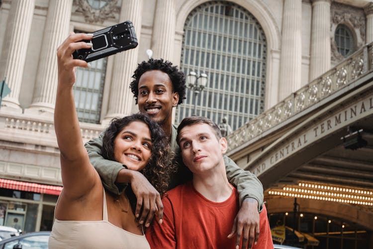 Positive Multiethnic Friends Taking Selfie On Modern Smartphone Near Railway Station Building