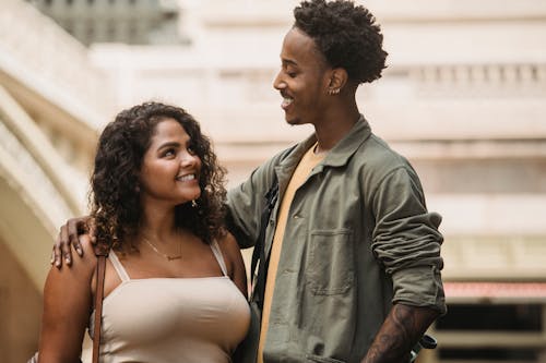 Positive multiracial young couple wearing casual clothes standing and hugging while looking at each other on blurred city street in daylight