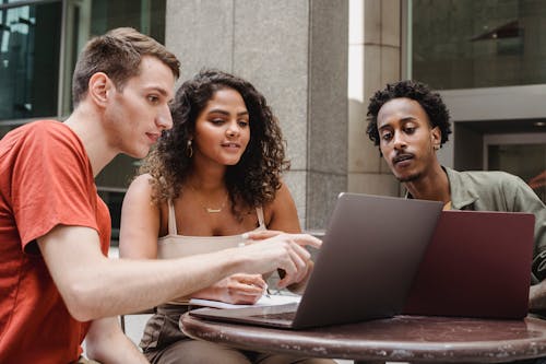 Free Group of diverse young male and female diverse colleagues discussing project details while working together on laptops in outdoors cafe Stock Photo