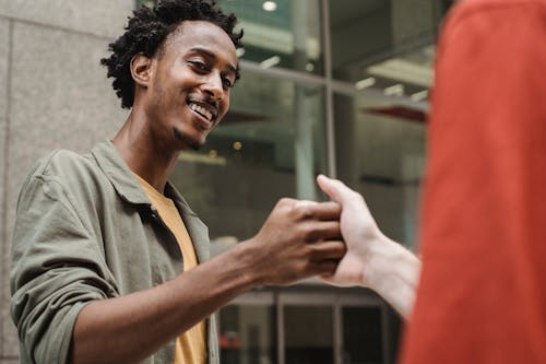 Low angle of cheerful young African American guy shaking hands with crop anonymous friend during meeting on city street