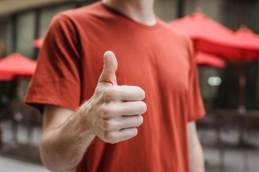 Crop anonymous young male in red t shirt showing thumb up gesture while standing on city street on sunny day