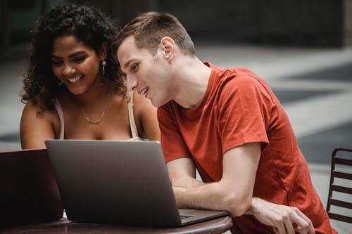 Joyful young multiracial couple in casual clothes smiling while sitting in outdoor cafe and studying online together using laptops