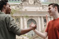 Side view of cheerful young multiracial male friends in trendy outfits smiling and looking at each other while shaking hands on city street after meeting