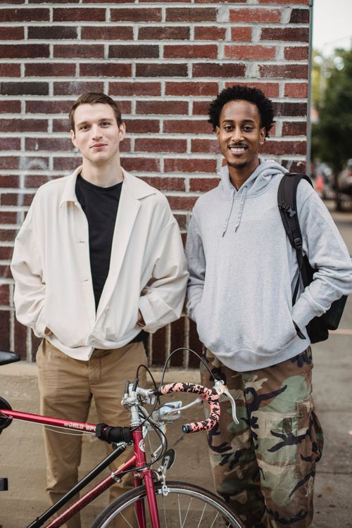 Free Cheerful young diverse male millennials in stylish outfits smiling and looking at camera while standing on street with hands in pockets near bicycle Stock Photo