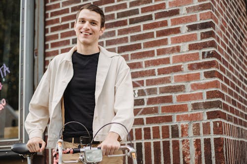 Low angle of cheerful young male millennial in stylish clothes smiling while standing on street with bicycle and looking at camera