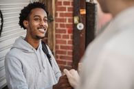Happy young African American man in stylish hoodie smiling while shaking hand of crop unrecognizable friends after meeting on city street