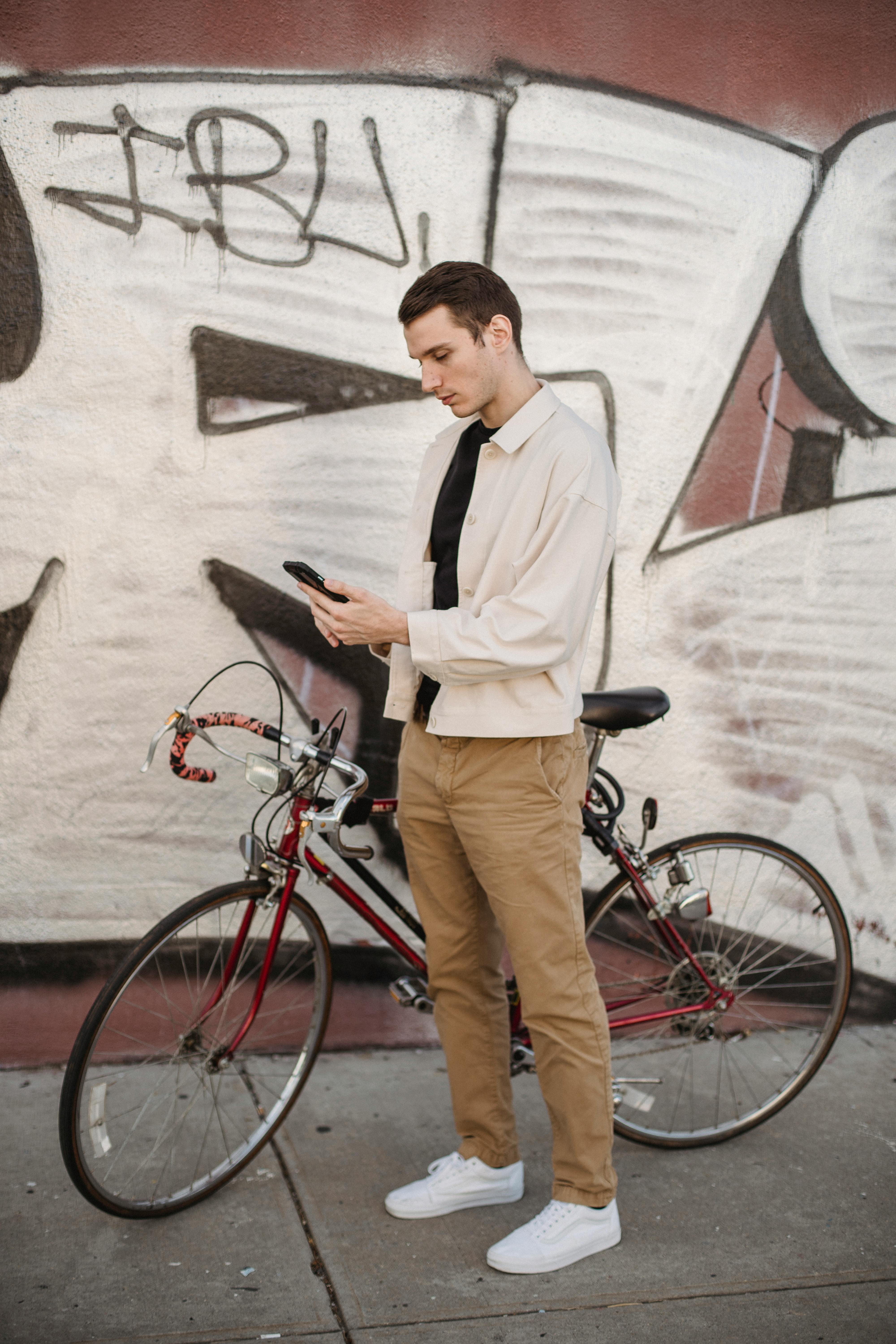 concentrated young guy surfing smartphone while standing near graffiti wall with bike