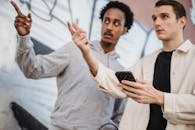 Serious young multiracial men pointing away and using smartphone near graffiti wall