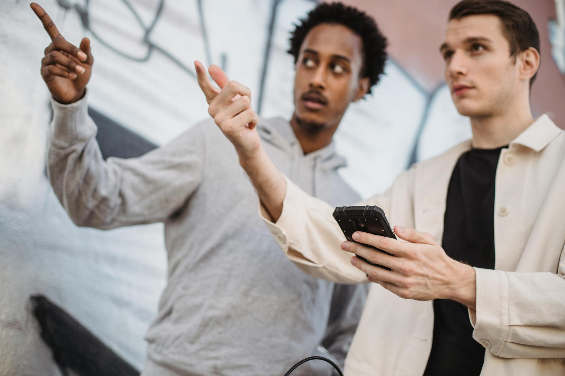 Two men pointing and using a smartphone to navigate directions outdoors in a casual setting.