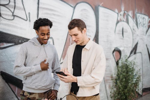 Positive young diverse male friends in trendy outfits smiling while watching video on smartphone standing near graffiti wall in city
