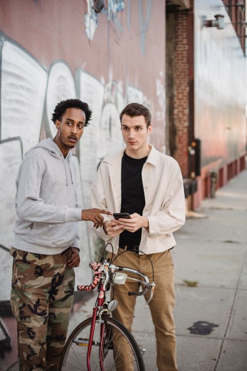Free Focused young multiethnic male travelers in casual outfits using online map on smartphone and looking away while standing on city street with bicycle near graffiti wall Stock Photo