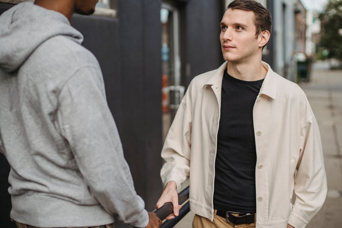 Free Serious young man in casual clothes standing on street and communicating with crop anonymous black male friend Stock Photo