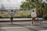 Back view of anonymous young male millennial in trendy outfit rolling bicycle while crossing asphalt road with African American friends in city outskirts