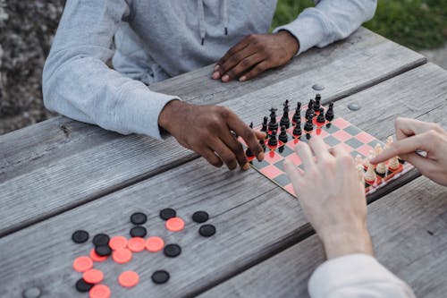 Free From above of crop unrecognizable multiethnic male friends sitting at wooden table in park and playing chess Stock Photo