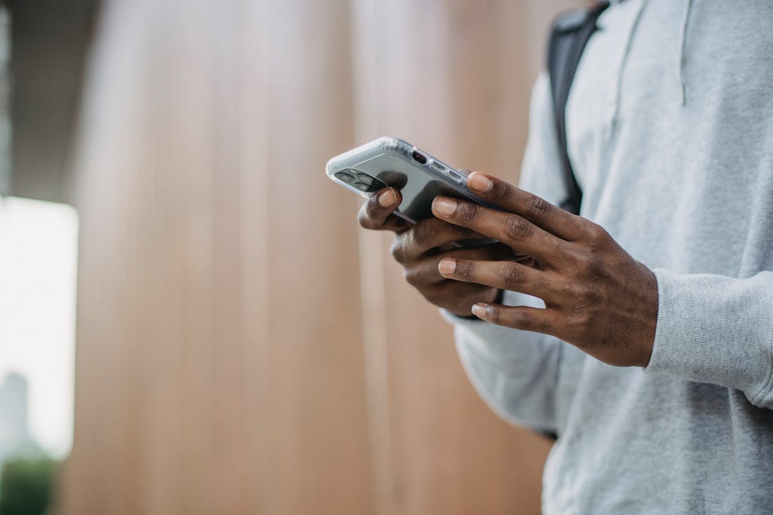 Free From below crop of unrecognizable African American young guy in casual clothes messaging on mobile phone while standing near wooden wall Stock Photo