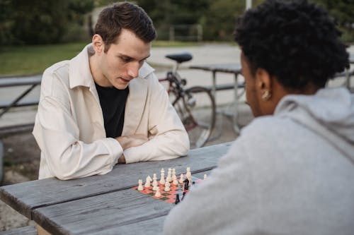 Concentrated young guy in casual clothes thinking while playing chess game with anonymous African American male friend in park