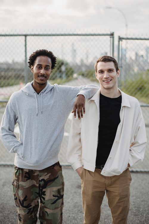 Free Smiling black man leaning on shoulder of positive young friend while standing against grid fence and looking at camera Stock Photo