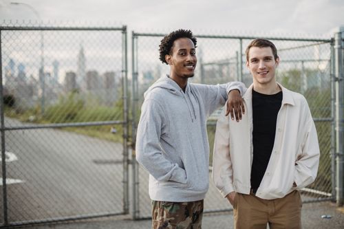 Free Positive multiethnic male friends wearing casual clothes standing close together against metal grid and smiling at camera Stock Photo