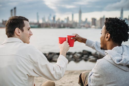Back view of diverse men clinking red cups sitting close on river bank and enjoying free time together