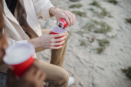High angle of crop anonymous male pouring refreshing drink into red cup while sitting on sandy shore with black friend