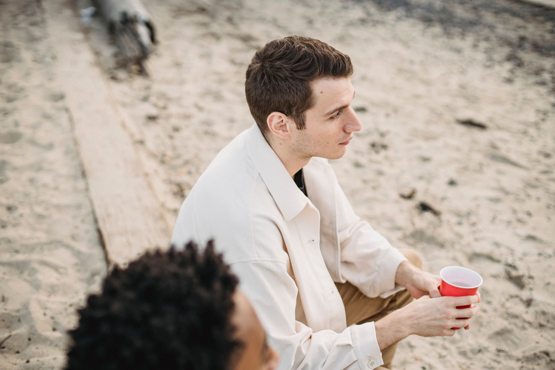 High angle of peaceful man with plastic cup sitting with African American friend on sandy coast