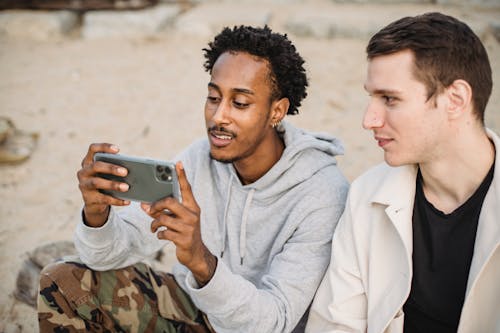 Free Attentive African American male sharing mobile phone with friend and watching video on sandy ground Stock Photo