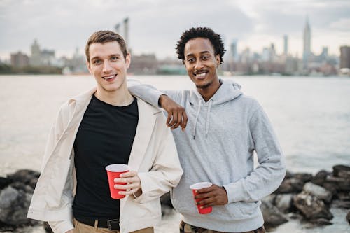 Free Positive diverse male friends wearing casual clothes standing close with red cups on river bank and looking at camera Stock Photo
