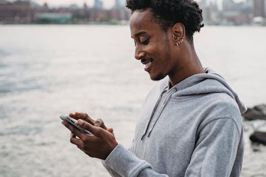 A young man enjoying his smartphone on a waterfront, smiling with a blurred city backdrop.