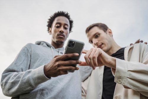 Free From below of serious black man hugging friend and scrolling mobile phone together against blurred cloudy sky Stock Photo