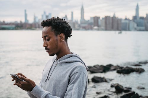 Side view of African American man scrolling mobile phone standing on coast of river against high buildings