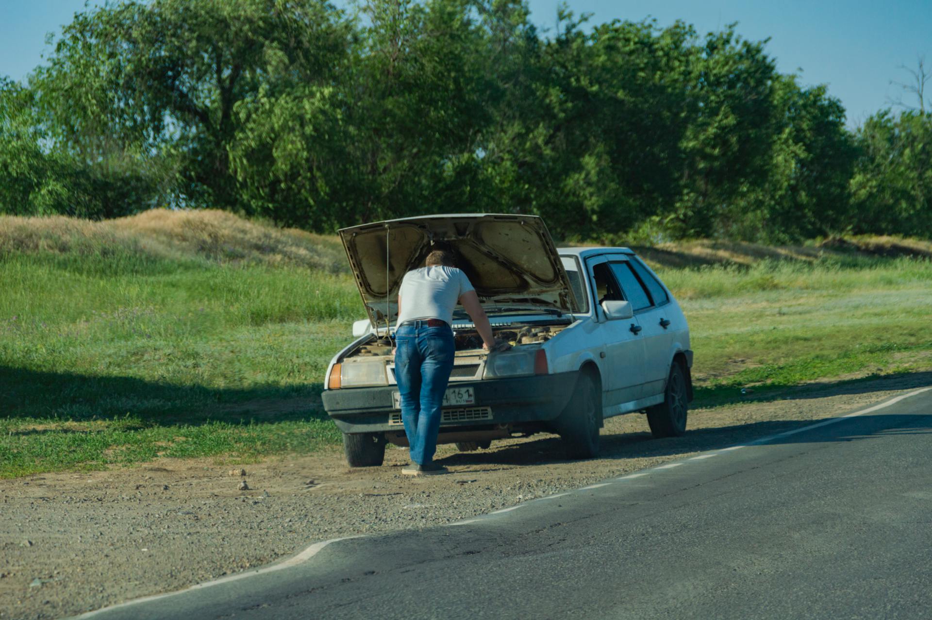 A man stands by his car on the roadside, inspecting a breakdown issue.