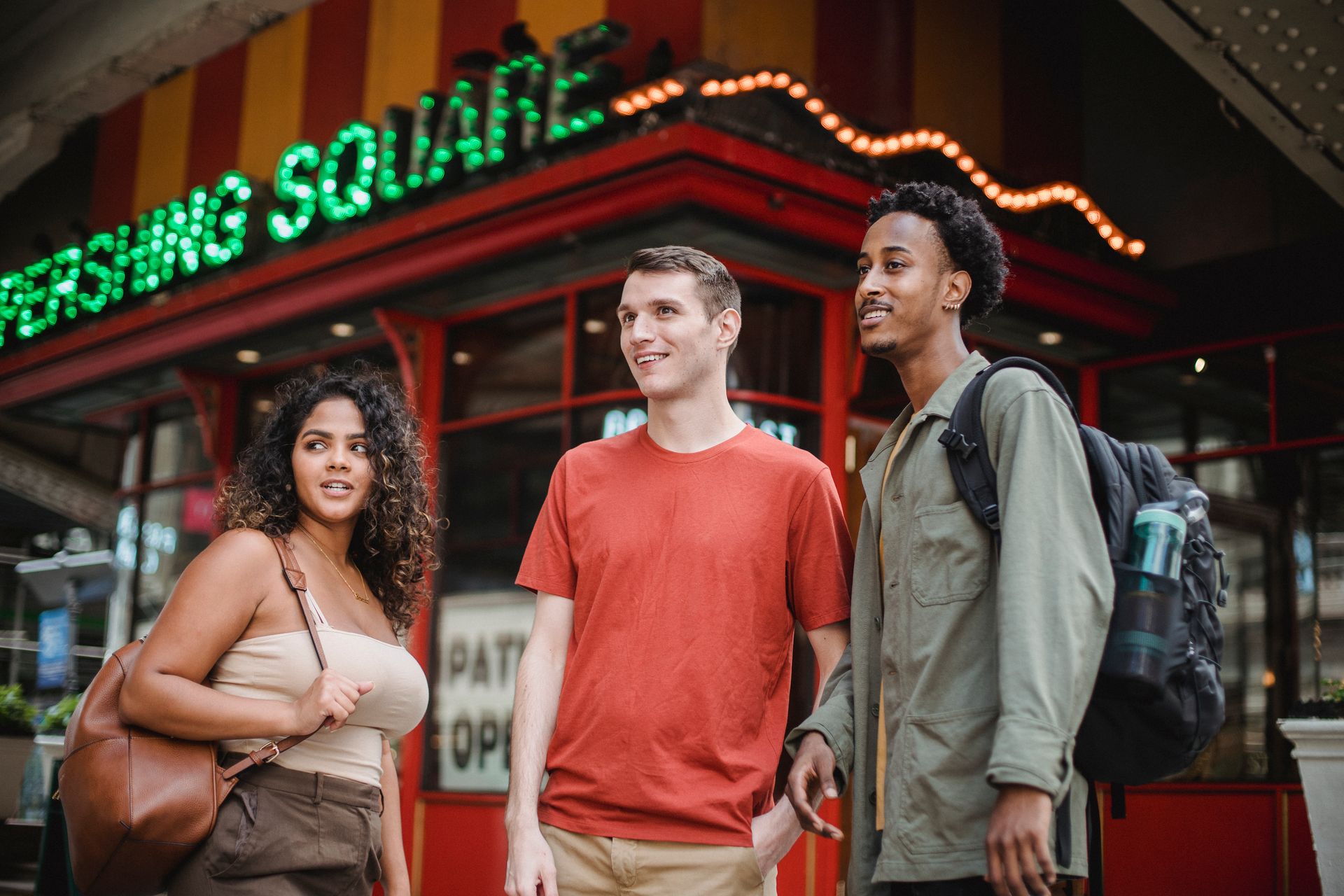 Group of young multiracial friends in casual outfits with backpacks standing in city street near building with sign and looking away in daytime