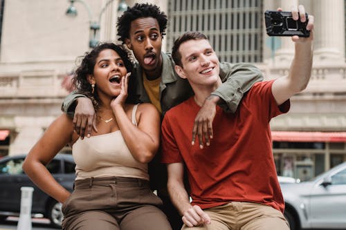 Positive young multiethnic friends in casual clothes taking selfie on phone while making funny faces in city street